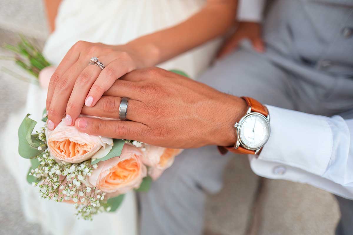 Photo des mains d’un couple nouvellement marié, montrant leurs alliances et un bouquet de roses pastel, symbole d’amour et d’engagement.
